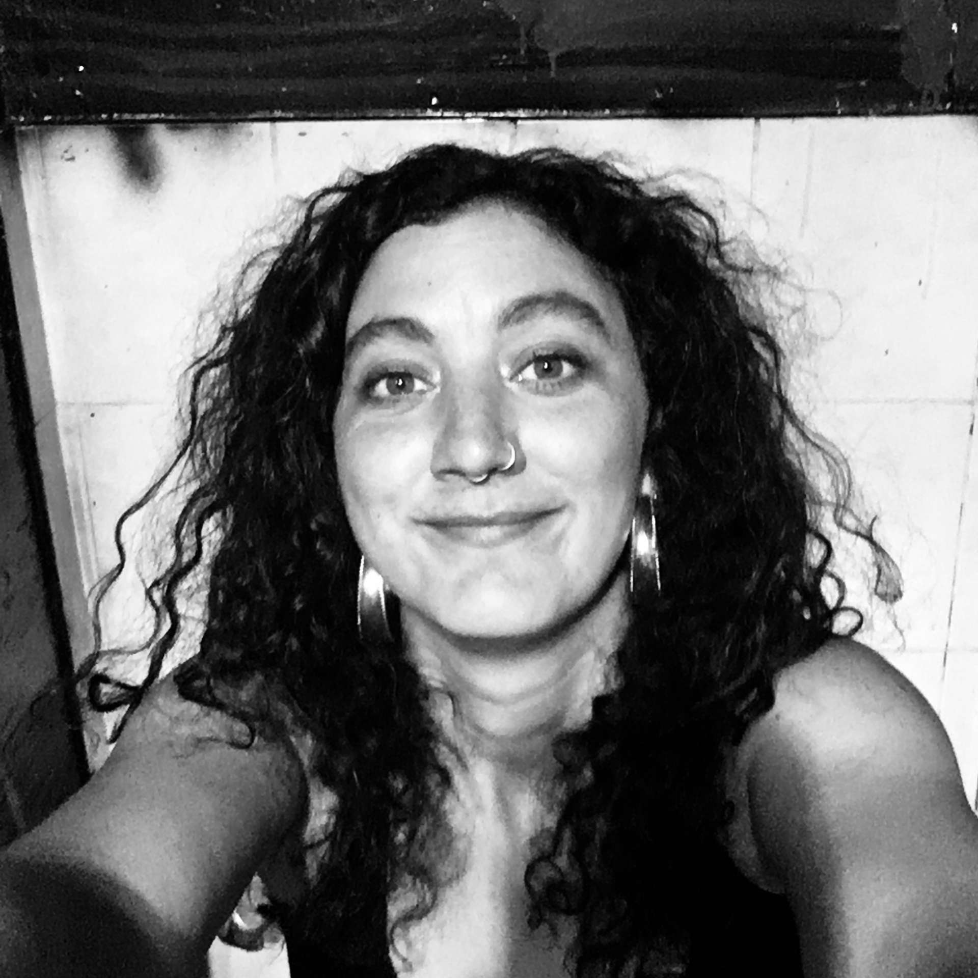 A black and white selfie of the writer, Mykaela Saunders. Mykaela is smiling at the camera and has long curly hair that falls over her shoulders.