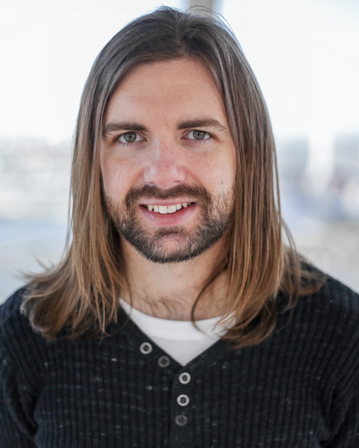 A headshot of the writer Beau Windon. Beau has long brown hair to his shoulders and a neat beard. Beau is wearing a dark coloured sweater over a white tshirt.